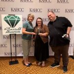 Three people holding an award at KAHCC Emerald Awards event with logo backdrop and sign reading “The Emerald Awards.”