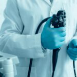 Clinician in blue gown and gloves holding endoscope controls beside medical equipment in clinical setting.