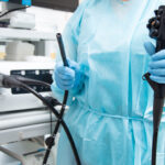 Clinician in blue gown and gloves holding endoscope controls beside medical equipment in clinical setting.